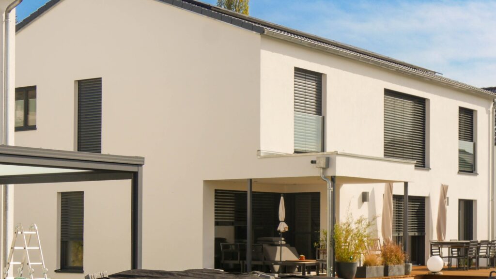 Modernes Einfamilienhaus mit weißer Putzfassade, Flachdach, Jalousien und überdachter Terrasse im Garten